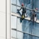 Two workers wearing safety gear are suspended by ropes on the side of a glass building, cleaning the windows. They are equipped with buckets and tools as they perform their task at significant heights.