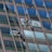 Two window cleaners are suspended by ropes, working on cleaning the glass windows of a tall building. Each cleaner is equipped with tools and a bucket, skillfully moving along the facade with safety harnesses. The building features reflective blue-tinted glass panels, creating a striking geometric pattern.