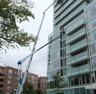 A glass-covered building is being cleaned or maintained using hydraulic boom lifts. Two platforms with workers are visible on the side of a high-rise structure. The scene is set in an urban environment with additional buildings visible in the background. Traffic cones and a small car are on the street below, suggesting maintenance work or caution.