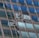 Two window cleaners are suspended by ropes, working on cleaning the glass windows of a tall building. Each cleaner is equipped with tools and a bucket, skillfully moving along the facade with safety harnesses. The building features reflective blue-tinted glass panels, creating a striking geometric pattern.