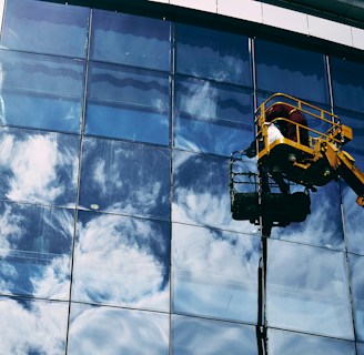 A worker in a yellow hydraulic lift platform cleans a large, reflective glass facade of a building. The sky with white clouds is vividly reflected in the mirrored surface of the windows. The sunlight casts bright highlights on the glass, enhancing the reflection of the clouds.
