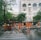 A group of workers wearing orange safety uniforms and helmets are cleaning a paved street area near a crosswalk. They are using hoses and brooms to clean the wet surface. Surrounding buildings, including a hotel named 'ROYAL HOTEL SAIGON,' and trees can be seen in the background.