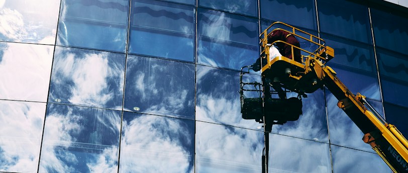 A worker in a yellow hydraulic lift platform cleans a large, reflective glass facade of a building. The sky with white clouds is vividly reflected in the mirrored surface of the windows. The sunlight casts bright highlights on the glass, enhancing the reflection of the clouds.