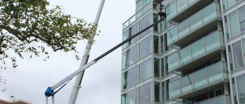 A glass-covered building is being cleaned or maintained using hydraulic boom lifts. Two platforms with workers are visible on the side of a high-rise structure. The scene is set in an urban environment with additional buildings visible in the background. Traffic cones and a small car are on the street below, suggesting maintenance work or caution.